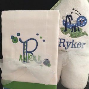 May include: A white hooded towel with a blue and green embroidered bug and the name "Ryker" on it. A white washcloth with a blue and green embroidered letter "R" and the name "Ryker" on it. The washcloth has a green and blue polka dot trim.