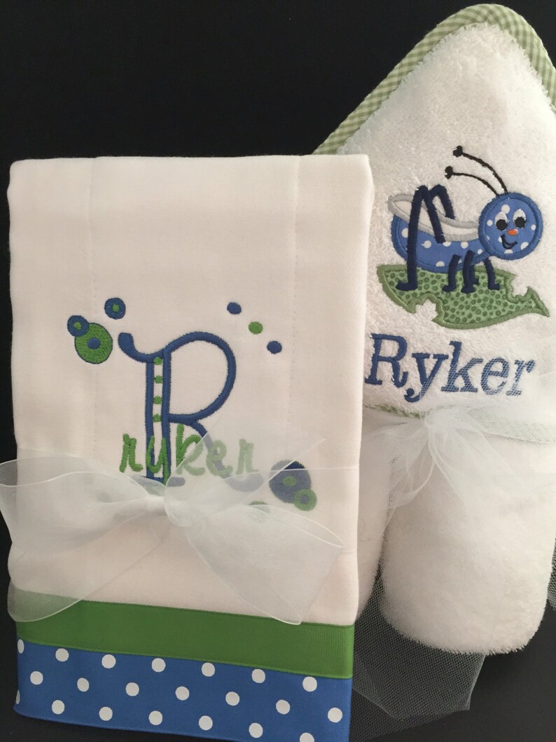 May include: A white hooded towel with a blue and green embroidered bug and the name "Ryker" on it. A white washcloth with a blue and green embroidered letter "R" and the name "Ryker" on it. The washcloth has a green and blue polka dot trim.