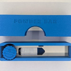May include: A bright blue "POWDER BAR" tool with a rectangular design. The top piece is separate from the bottom, which has a dial and a silver blade. The tool is designed for a specific purpose.