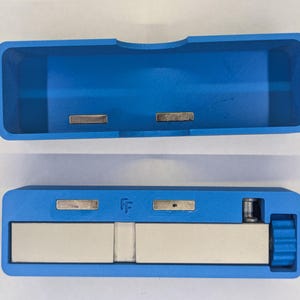 May include: A blue rectangular case with a matching lid, both featuring embedded silver-colored metal components. The case is open, revealing a larger, rectangular silver-colored piece inside. The design is modern and functional.