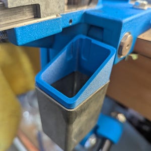 May include: Close-up of a reloading press with a bright blue frame and a silver metal component. A small, rectangular, dark gray catch tray with a blue rim is attached to the press.