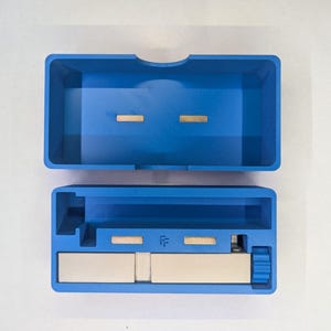 May include: A blue plastic box with a lid, open to reveal the interior. The box has two small, rectangular gold-colored metal pieces inside. The bottom section has a silver-colored metal piece and a small blue plastic piece.