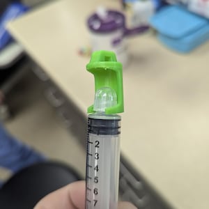 May include: A clear plastic syringe with black measurement markings from 2 to 10. A green plastic cap is attached to the top. The syringe is held in a hand.