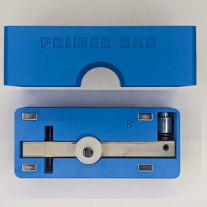 Op de afbeelding: Een blauwe primer bar met de tekst "PRIMER BAR" op het deksel. De open bar onthult een zilveren metalen mechanisme met een wit cirkelvormig onderdeel. De bar is ontworpen voor het herladen van munitie.