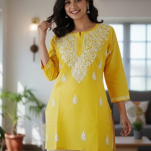 May include: A yellow tunic with white floral embroidery and three-quarter sleeves. The tunic is paired with white palazzo pants. The outfit is a bright, casual style, suitable for warm weather. The embroidery is detailed and adds a decorative element.
