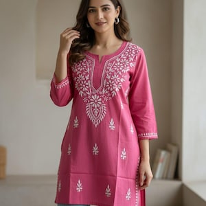 May include: A pink tunic with white floral embroidery. The tunic has a round neckline with a small slit and three-quarter sleeves. The embroidery is concentrated around the neckline and down the front, with smaller floral accents scattered across the garment.