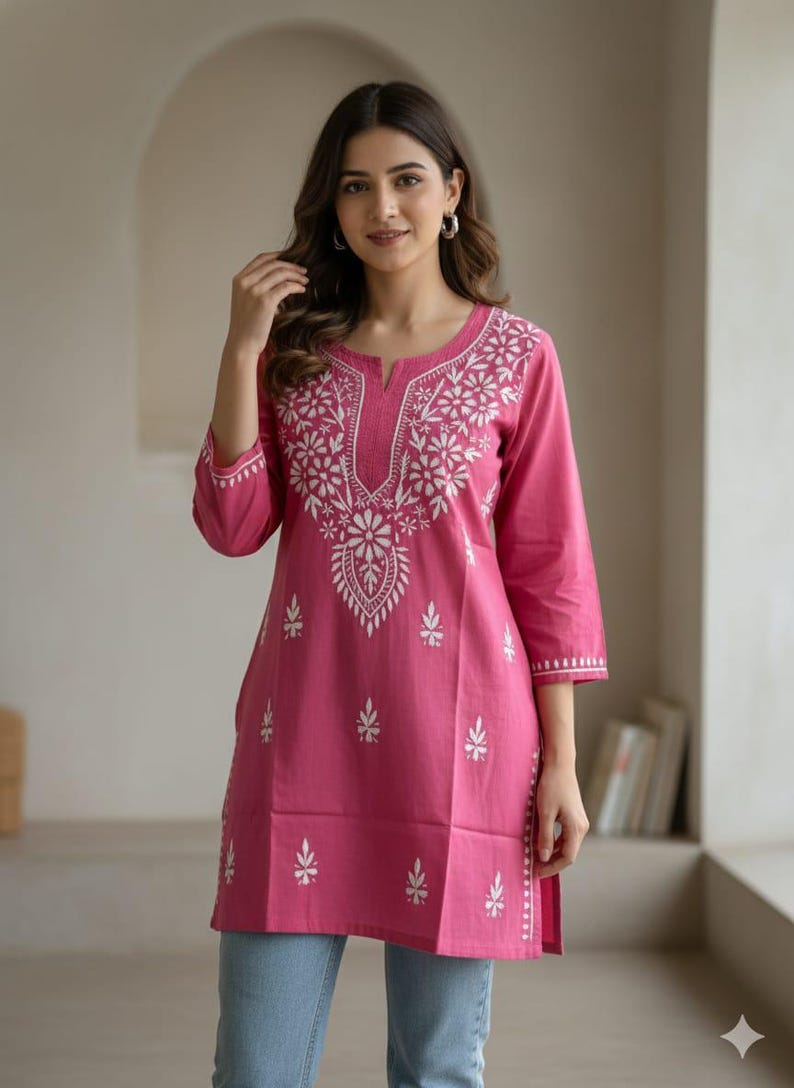 May include: A pink tunic with white floral embroidery. The tunic has a round neckline with a small slit and three-quarter sleeves. The embroidery is concentrated around the neckline and down the front, with smaller floral accents scattered across the garment.