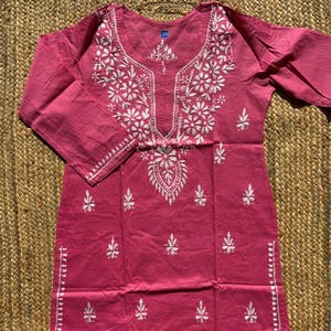 May include: A dusty rose tunic with white floral embroidery. The tunic features a round neckline and three-quarter sleeves. The embroidery is concentrated around the neckline and down the front, with smaller floral accents scattered across the garment.