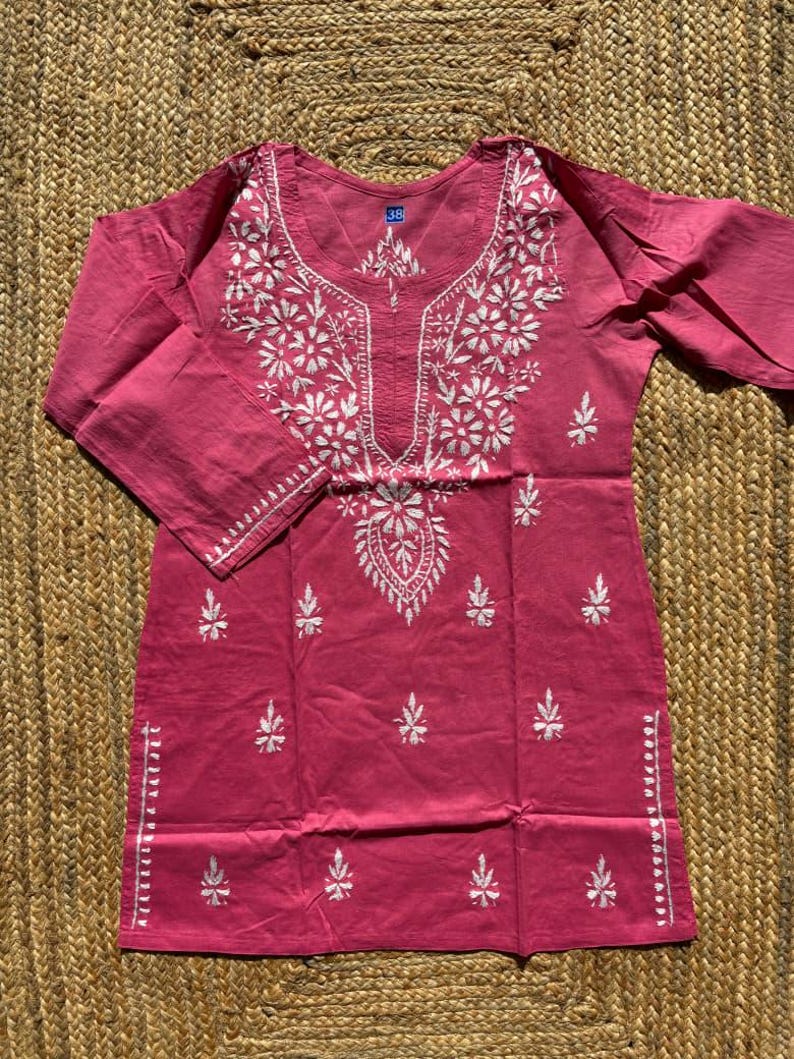May include: A dusty rose tunic with white floral embroidery. The tunic features a round neckline and three-quarter sleeves. The embroidery is concentrated around the neckline and down the front, with smaller floral accents scattered across the garment.