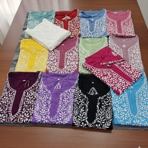May include: A collection of embroidered cotton tunics in various colors, including teal, red, blue, and purple. Each tunic features intricate white floral and leaf patterns around the neckline and sleeves. The garments are neatly folded and arranged on a table.