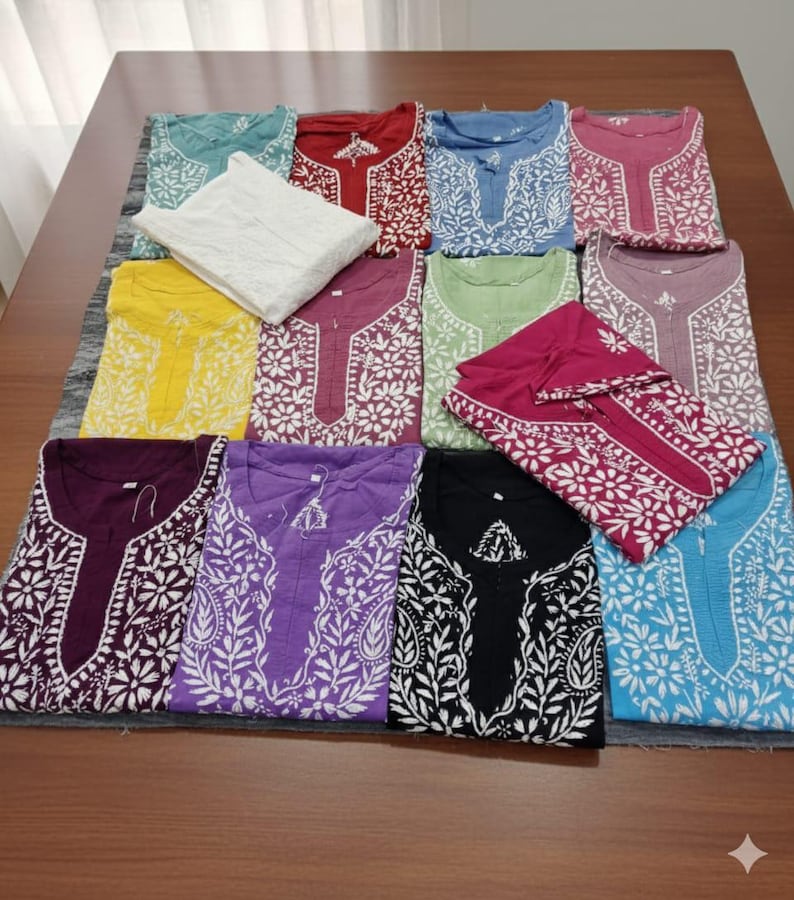 May include: A collection of embroidered cotton tunics in various colors, including teal, red, blue, and purple. Each tunic features intricate white floral and leaf patterns around the neckline and sleeves. The garments are neatly folded and arranged on a table.