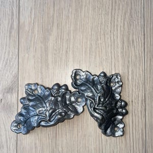 Könnte beinhalten: Zwei dekorative Metallverzierungen in Form von Eichenblättern und Eicheln. Die Blätter sind mit Adern und kleinen Löchern zur Befestigung versehen. Die Oberfläche ist ein dunkles, metallisches Grau mit Glanzlichtern.