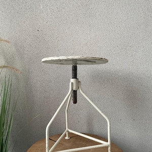 Pode incluir: Banqueta ajustável em estilo vintage com assento redondo branco sujo e estrutura metálica branca. O assento é suportado por uma haste roscada para ajuste de altura. A banqueta tem base de tripé.
