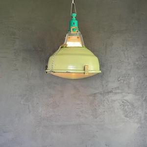 May include: A pendant light fixture featuring a large, pale green, dome-shaped metal shade. The top section is a bright teal color with a metal chain for suspension. A small metal latch is visible on the side of the shade, and the light is illuminated.