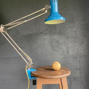 May include: A blue desk lamp with a silver adjustable arm is clamped to a wooden table. A baseball sits on the table. The lamp has a yellow cord and a blue lampshade with a yellow interior.