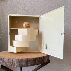 Pode incluir: Um armário de metal branco sujo com uma porta aberta, revelando uma bola de beisebol em uma prateleira e quatro gavetas claras descendentes. O armário está sobre uma mesa de madeira rústica com base de metal.