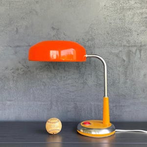 May include: A retro orange desk lamp with a curved chrome arm and a round base. A baseball sits to the left of the lamp. The lamp has a red button and a white cord.