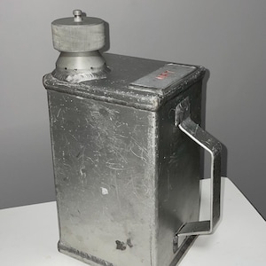 Puede incluir: Un contenedor de metal plateado con asa y tapón de rosca. El contenedor rectangular tiene un asa en un lado y un tapón en la parte superior. El metal tiene arañazos y desgaste visibles. El tapón es cilíndrico y tiene una textura acanalada.