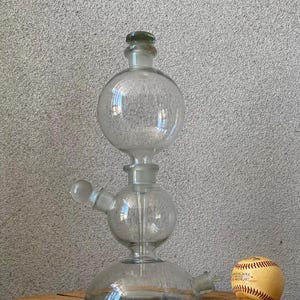 May include: A clear glass scientific apparatus with a bulbous design, featuring multiple spherical chambers and a flared base. A baseball with red stitching rests beside it. The apparatus is likely used for laboratory experiments or decorative purposes.