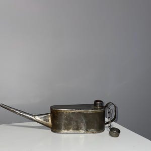 Puede incluir: Una aceitera de metal vintage con un pico largo y una pequeña tapa sujeta por una cadena. La lata tiene un cuerpo rectangular y un cuello de llenado cilíndrico. El metal tiene un aspecto deslustrado y envejecido.