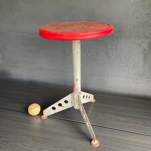 May include: A round stool with a red padded seat and a light grey metal base featuring three legs with circular cutouts. The metal base shows signs of wear and paint chipping. A yellow baseball is positioned on the floor near the stool base.