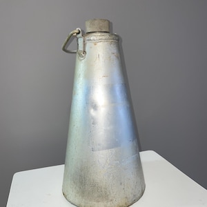 Puede incluir: Una cantimplora de metal plateado con forma cónica. Tiene una tapa de rosca y un pequeño lazo de metal para llevarla. La cantimplora está sobre una superficie blanca. La altura de la cantimplora es de unos 25 cm.