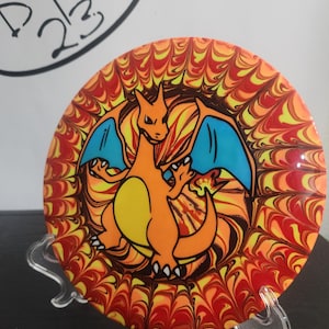 May include: A colorful disc golf disc featuring a Charizard design. The disc has a vibrant orange, yellow, and red pattern with a blue-winged Charizard in the center. The text "DYE 23" is visible in the upper left corner.