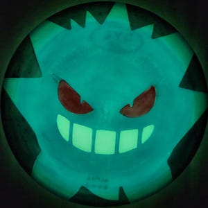 May include: A round, glow-in-the-dark disc golf disc featuring a luminous teal surface with a dark, stylized graphic of a grinning creature. The design includes two sharp, dark eyes and a wide, toothy smile, with dark triangular shapes along the edge.