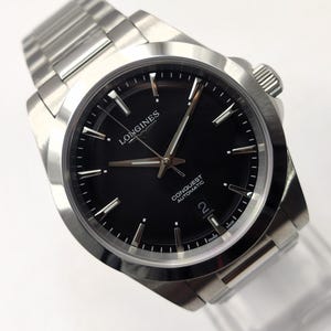 May include: A stainless steel Longines Conquest Automatic watch with a black dial, silver-tone hands, and hour markers. The watch features a date window at the six o'clock position, a brushed metal link bracelet, and the text Longines Conquest Automatic.