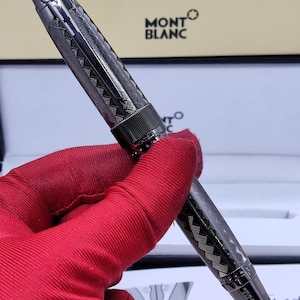 Könnte beinhalten: Ein dunkelgrauer Montblanc-Stift mit einem Muster. Der Stift hat ein strukturiertes Muster und einen silbernen Clip. Das Montblanc-Logo ist im Hintergrund sichtbar. Der Stift wird von einer Person mit rotem Handschuh gehalten.