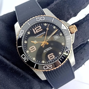 May include: A Longines automatic watch with a black dial, gold-tone hands and numerals, and a date window at the three o'clock position. The watch features a black ceramic rotating bezel with gold-tone accents, a stainless steel case, and a black textured rubber strap.