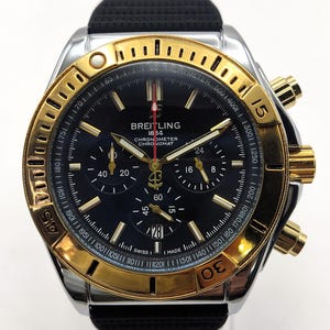 May include: A Breitling Chronomat watch with a black dial, gold-tone bezel, and silver-tone case. The watch features a black textured rubber strap, three sub-dials, a date window at the six o'clock position, and a red-tipped second hand.