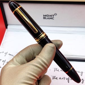 May include: A black Montblanc fountain pen with gold-tone accents, including a clip and rings on the cap. The pen features a glossy finish and is presented in a branded box with the text Montblanc visible on the interior lid and packaging.