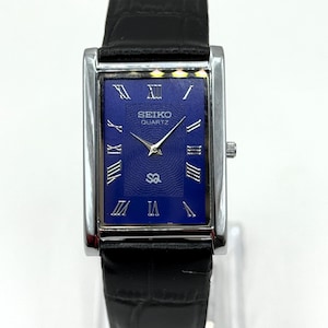 Könnte beinhalten: Eine rechteckige Seiko Quartz-Uhr mit blauem Zifferblatt und silbernen römischen Ziffern. Die Uhr hat ein silbernes Gehäuse und ein schwarzes Lederarmband. Das Zifferblatt zeigt das Seiko-Logo und das Wort "Quartz".