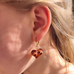 Puede incluir: Un pendiente en forma de corazón con un gancho dorado. El corazón es marrón translúcido con manchas marrones más oscuras. El pendiente cuelga de una oreja.