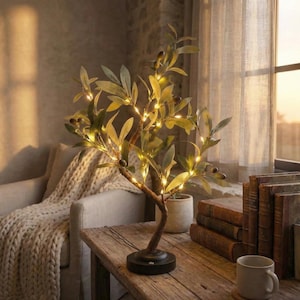 Könnte beinhalten: Dekorative Olivenbaumlampe mit warmweißen LED-Lichtern. Die Lampe hat einen braunen Stamm, grüne Blätter und kleine schwarze Olivenakzente. Sie steht auf einem schwarzen Sockel auf einem rustikalen Holztisch.