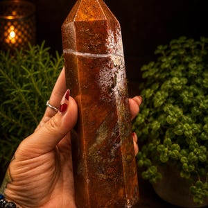 May include: A polished, hexagonal crystal tower featuring a deep reddish-brown base color with intricate, moss-like white and dark green inclusions throughout the stone, culminating in a sharp, pointed apex at the top of the vertical structure.