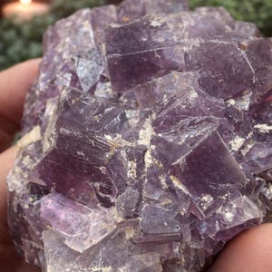 Peut inclure: Gros plan sur un amas de cristaux de fluorite violets. Le cristal a une structure cubique avec diff&eacute;rentes nuances de violet et de blanc. L'arri&egrave;re-plan est flou, avec une bougie et de la verdure visibles.
