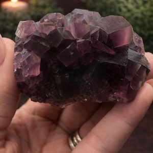 Peut inclure: Une main tient un grand cristal de fluorite violet facett&eacute;. Le cristal a une couleur riche et profonde avec des variations de tons. L'arri&egrave;re-plan est flou, avec une bougie et de la verdure visibles. Cet objet est d&eacute;coratif.