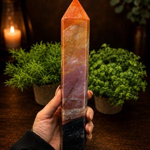 May include: A tall, multi-colored crystal tower with a pointed top. The stone features a gradient of colors, transitioning from a bright orange at the apex through shades of tan, deep red, and brown, ending in a solid black base with white veining.