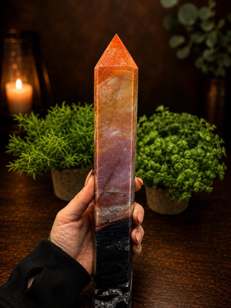 May include: A tall, multi-colored crystal tower with a pointed top. The stone features a gradient of colors, transitioning from a bright orange at the apex through shades of tan, deep red, and brown, ending in a solid black base with white veining.