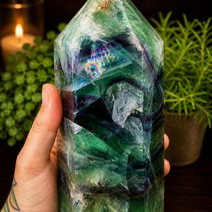 May include: A large, faceted fluorite crystal tower with shades of green, blue, and purple. The crystal is held in a hand and has a polished, reflective surface. The crystal is approximately 6 inches tall.