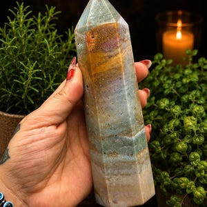 May include: A tall, hexagonal crystal tower featuring a polished surface with layered bands of muted blue, earthy brown, and soft ochre tones. The stone displays natural mineral inclusions and a smooth, faceted finish that tapers to a point.