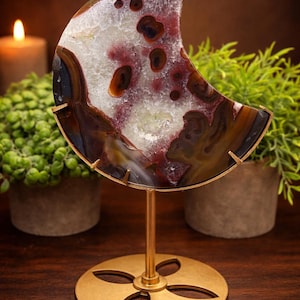 May include: A crescent-shaped agate slice, showcasing a mix of colors including white, burgundy, and brown, mounted on a gold-colored metal stand. The stand has a circular base with a floral design. Decorative item.