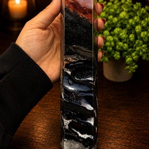May include: A tall, multi-faceted stone tower featuring a distinct color gradient. The top section displays a deep reddish-brown hue, transitioning into a solid black middle, and ending in a base with intricate black and white banded agate patterns.