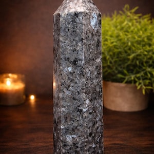 May include: A grey granite obelisk with a pointed top. The stone has a speckled pattern of black and silver. The obelisk is standing upright on a wooden surface. A small potted plant and a lit candle are in the background.