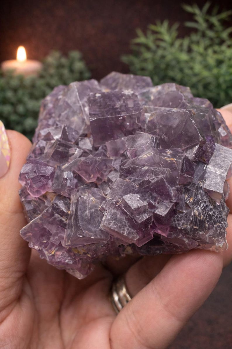Peut inclure: Un grand amas de cristaux de fluorite violette brute. Le cristal pr&eacute;sente une structure g&eacute;om&eacute;trique complexe avec de nombreuses facettes et une gamme de teintes violettes. L'arri&egrave;re-plan comprend une bougie allum&eacute;e et de la verdure.