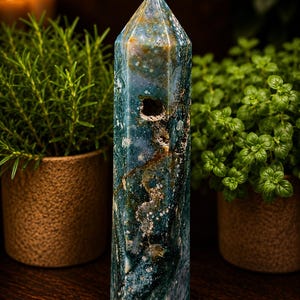 May include: A polished hexagonal crystal tower featuring a deep teal and forest green base with intricate white and brown mineral inclusions. The stone displays a natural textured cavity on one side, with a smooth, pointed top and faceted sides.