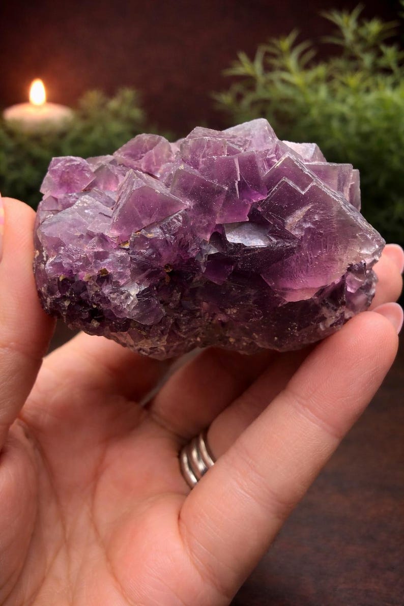 Peut inclure: Une main tient un cristal de fluorite brut et naturel. Le cristal est de couleur violet fonc&eacute; avec une structure cristalline. L'arri&egrave;re-plan est flou, avec une bougie allum&eacute;e et de la verdure visible.