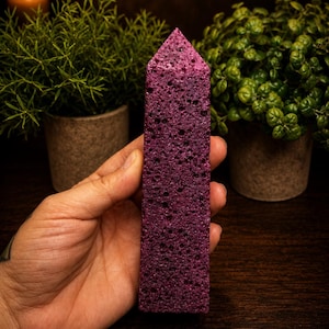 May include: A tall, six-sided obelisk-shaped crystal tower with a deep magenta, porous, and textured surface. The stone features a pointed top and a rough, volcanic-like appearance with numerous small, dark indentations across its entire body.